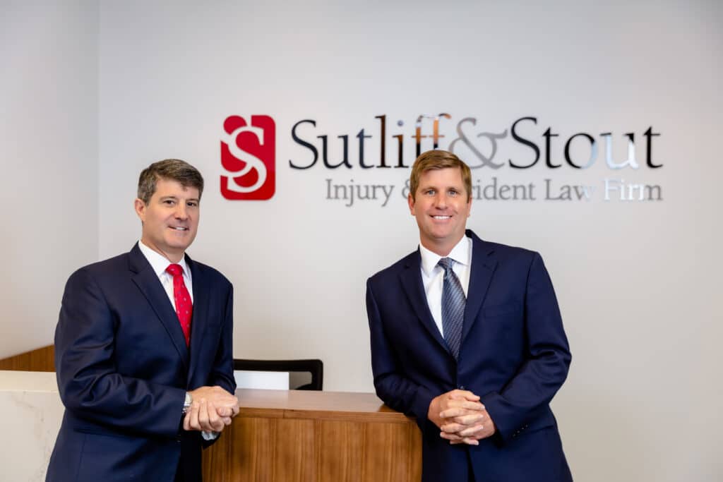 Graham Sutliff and Hank Stout stand at the entrance to their Austin based law firm. A sign that reads Sutliff & Stout Personal Injury Law Firm can be read in the background.