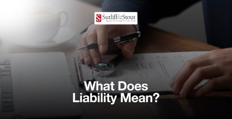 Person's hands holding pen over legal documents at desk with Sutliff & Stout logo and "What Does Liability Mean?" text